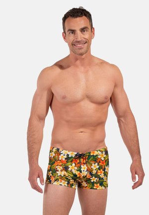 Smiling muscular man wearing floral patterned swim trunks with orange, white flowers and green leaves on light background.