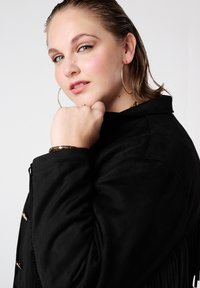 Zwarte suède jas met franjes, voorzien van zilverkleurige hardware. Soepele textuur met een gestructureerde kraag. Model toont een casual houding.