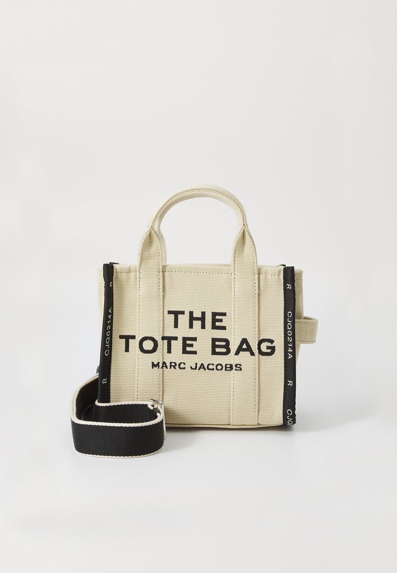Saco de tela bege com alças curtas, texto preto "THE TOTE BAG MARC JACOBS," e alça de ombro preta destacável com bordas brancas.