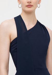 Vestido azul-marinho com uma alça única e detalhes em camadas de tule, apresentando um corpete ajustado e textura suave. Brincos de argola prateados incluídos.