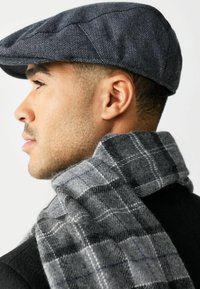Man in profile wearing a dark grey herringbone flat cap and a thick grey plaid scarf over a black coat against a white background.