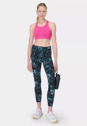 Vrouw staand in bloemenleggings en roze sportbeha, houdend een zwarte waterfles, draagt witte sneakers tegen een witte achtergrond.