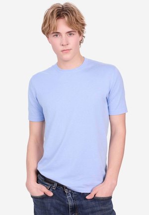 Junger Mann mit blondem Haar, der ein hellblaues Kurzarmhemd und dunkle Jeans trägt, steht mit den Händen in den Taschen vor einfachem Hintergrund.