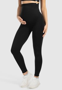 Leggings de maternité noirs avec une taille haute, texture lisse et design longueur entière. Portés avec des chaussures de sport blanches.