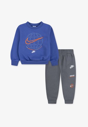Nike dviejų dalių komplektas, kurį sudaro mėlynas džemperis su pasaulio grafika ir oranžiniais akcentais, derinamas su pilkais sportiniais džinsais su logotipo detalėmis.