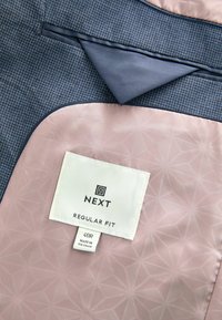 Puvun takin vuori, jossa on vaaleanpunainen geometrinen kuvio, ja jossa on "NEXT Regular Fit 40R Made in Vietnam" -merkki, sekä näkyvissä sininen ulkopinta.