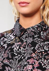 Schwarze Bluse mit floralem Muster und Kragen. Verfügt über komplexe weiße und farbige Blumenmuster. Das Material hat eine glatte Textur.