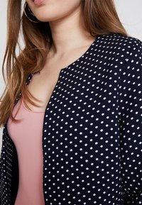 Blazer en tissu marine avec des pois blancs, un col rond et une coupe ajustée. Texture lisse avec un léger éclat.