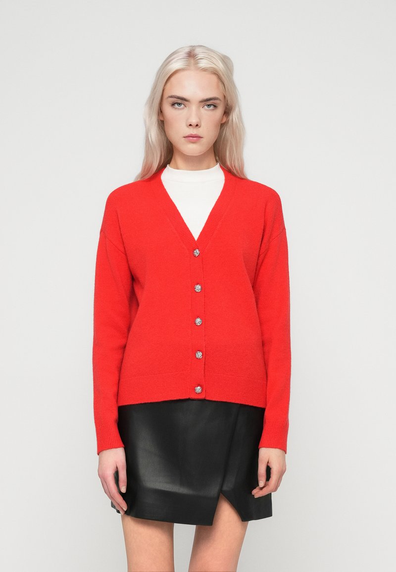 Mulher loira vestindo um cardigã vermelho brilhante abotoado sobre uma blusa branca, combinado com uma saia de couro preta, estando em frente a um fundo simples.