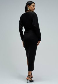 Vestido de punto negro con mangas largas, diseño ajustado, abertura trasera y escote redondo. Complementado con tacones altos negros con tiras.