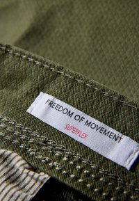 Tkanina w oliwkowej zieleni z widocznym przeszyciem, z białą etykietą, na której czerwonymi literami napisano "FREEDOM OF MOVEMENT" oraz "SUPERFLEX".