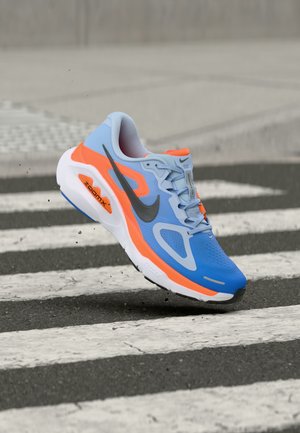 Chaussure de course Nike ZoomX bleue et orange en l'air au-dessus d'un passage pour piétons dans une rue, avec de petites particules de terre sous la semelle.