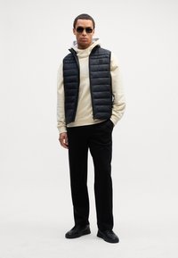 Μαύρο γιλέκο με πούπουλα πάνω από ένα ανοιχτό μπεζ φούτερ, συνδυασμένο με μαύρο παντελόνι και μαύρα αθλητικά παπούτσια. Μοντέλο που φορά γυαλιά ηλίου και στέκεται μπροστά σε έναν ουδέτερο φόντο.