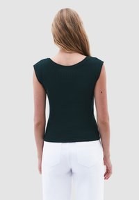 Maglia corta a maniche corte di un verde scuro a coste con un ampio scollo, abbinata a pantaloni bianchi a vita alta. Il tessuto ha una trama liscia.