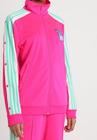 Chaqueta de chándal Adidas de color rosa brillante con mangas a rayas en tonos verde azulado y blanco, detalles de botones y bolsillos frontales, llevada por una persona.