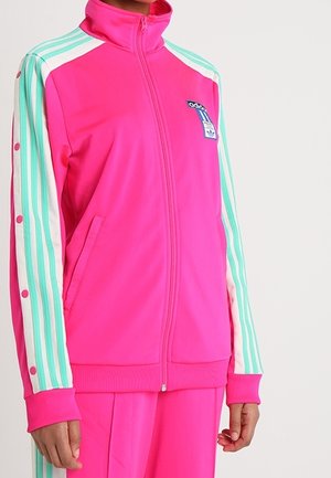 Chaqueta de chándal Adidas de color rosa brillante con mangas a rayas en tonos verde azulado y blanco, detalles de botones y bolsillos frontales, llevada por una persona.