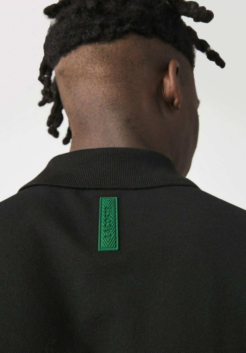 T-shirt polo noir avec un col uni et une patch texturée verte au dos avec un design en relief. Texture de tissu lisse.