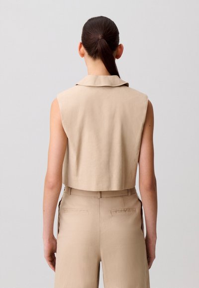 Donna con capelli scuri legati in una coda di cavallo, indossa un giubbotto beige senza maniche corto e pantaloni beige a vita alta coordinati, vista da dietro.