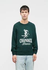 Temnozelen pulover iz pletene tkanine z grafiko smučanja in besedilom "CHAMONIX Francija" v belem. Rebrast krojeni ovratnik in manšete.