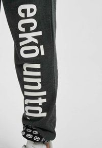 Mørkegrå sweatpants med store hvide bogstaver "eckō unlt'd" langs siden, med en afsluttet kant og et mønstret indre.