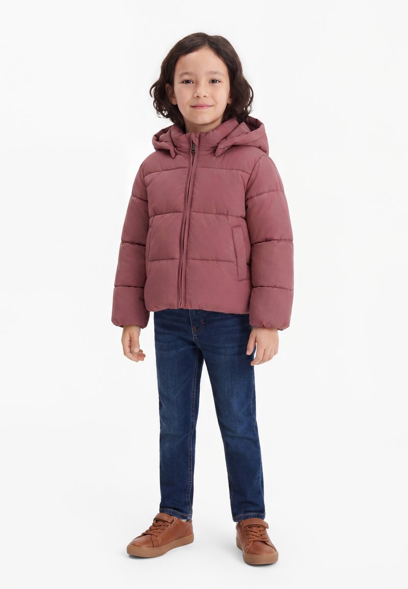 Enfant aux longs cheveux foncés portant une doudoune mauve, un jean bleu et des baskets marron, debout avec un léger sourire.