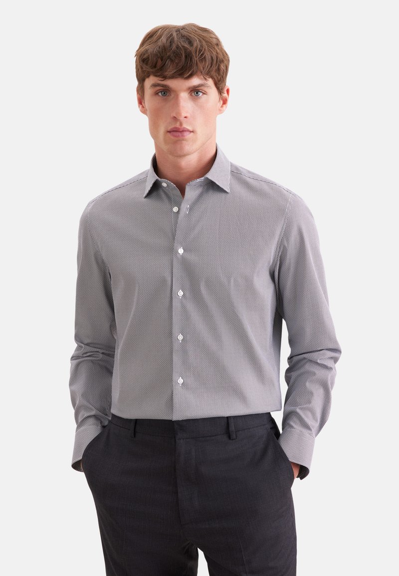 Jeune homme portant une chemise à carreaux slim fit à manches longues rentrée dans un pantalon foncé, les mains dans les poches, faisant face vers l’avant.