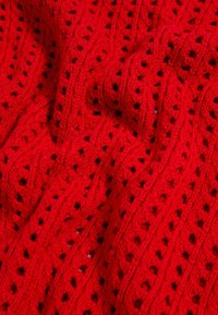 Roter Strickstoff mit einem strukturierten Wellenmuster und rautenförmigen Ausschnitten, der ein dickes Garn mit einer glatten Oberfläche zeigt.