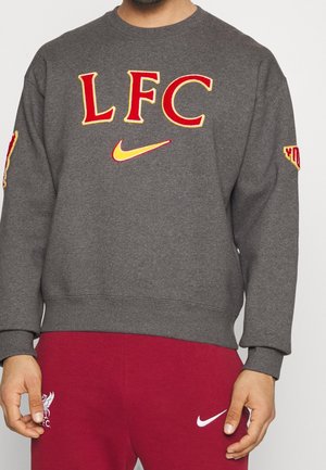 Personne portant un sweat-shirt gris avec les logos rouge et jaune "LFC" et Nike, associé à un pantalon rouge avec les logos Liverpool FC et Nike.