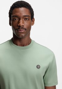 Light green short-sleeve T-shirt in smooth fabric with a round neckline, featuring a small round black logo patch on the left chest.