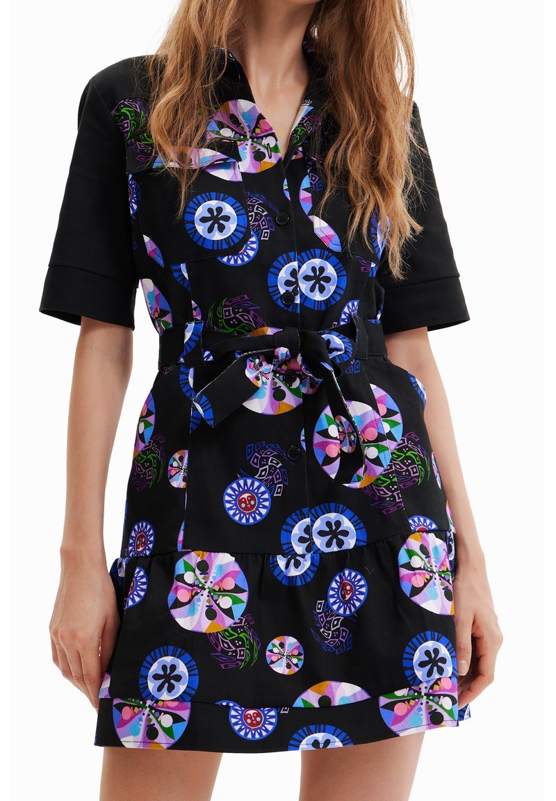 Femme portant une robe noire à manches courtes avec des motifs abstraits circulaires colorés, cintrée à la taille par une ceinture en tissu.