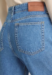 Close-up van de achterzak van blauwe denim jeans met een bruine Marc O’Polo merklabel gestikt op de tailleband.