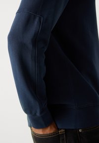 Felpa blu navy con collo a giro, polsini a costine e texture liscia. Dettagli delle cuciture visibili lungo la manica. Vestibilità casual.