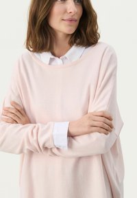 Maglione a maglia di un rosa chiaro con vestibilità ampia, scollatura rotonda e maniche lunghe. Indossato sopra una camicia bianca con colletto e dettagli dei bottoni.