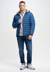 Niebieska kurtka puchowa z żółtym kapturem, z poziomymi przeszyciami. Noszona na szarym swetrze, w połączeniu z niebieskimi dżinsami i szarymi sneakersami.