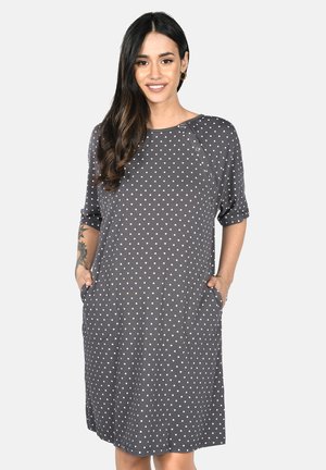 Woman with long dark hair wearing a grey knee-length dress with white heart patterns and hands in pockets, smiling at the camera.