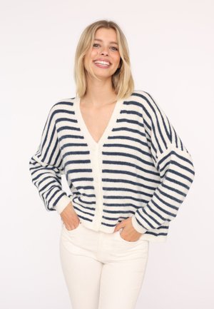 Lächelnde Frau mit blondem Haar, die eine weiße und marineblaue gestreifte Strickjacke mit Knöpfen und weiße Hosen trägt, Hände in den Taschen.