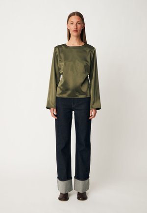 Femme debout face à nous, portant un haut vert en satin à manches longues, un jean foncé retroussé et des bottines marron sur un fond uni.