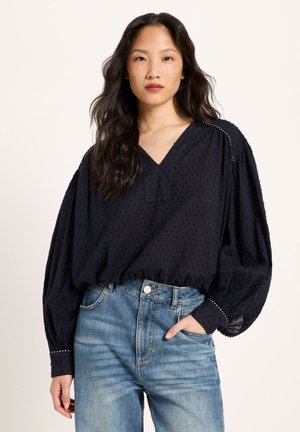 Frau mit langen schwarzen Haaren, die eine lockere schwarze strukturierte Bluse mit Ballonärmeln und blaue hoch taillierte Jeans trägt, vor einem schlichten Hintergrund stehend.