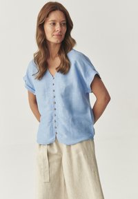 Lysblå linnedbluse med korte ærmer, V-hals, knaplukning foran og buet kant. Sammen med kridhvide, vide bukser.