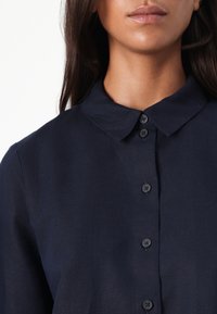 Mørkeblå skjorte med krage, laget av teksturert stoff og med seks svarte knapper foran.