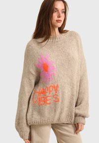 Pull en tricot beige à coupe surdimensionnée, avec une fleur rose vif et un texte orange "HAPPY VIBES". Poignets et ourlet côtelés.