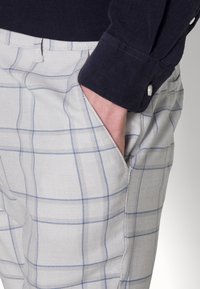 Ljusgrå rutiga byxor med blå ränder. Tillverkade av vävt tyg, med sidofickor och sydda detaljer. Marinblå långärmad skjorta synlig.
