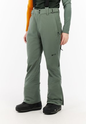 Persona con pantalones de esquí verdes con tirantes y botas negras de nieve de pie sobre un fondo blanco.
