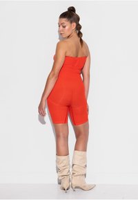 Moteris su pynėmis, vilkinti oranžinį be petnešėlių kombinezoną ir smėlio spalvos laisvalaikio aukštakulnius, stovinti ir žvelgianti per petį.