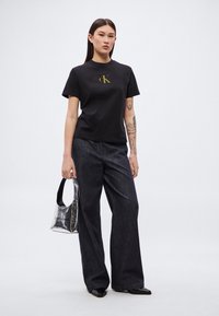 Personne portant un t-shirt noir Calvin Klein, un jean large sombre, des chaussures pointues noires, tenant un petit sac à main métallique Calvin Klein, avec des tatouages sur le bras gauche.