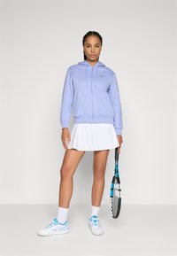 Lyselilla zip-up hettegenser med snorer, hvit plisserte skjørt, hvite joggesko med blå detaljer, holder en tennisracket.