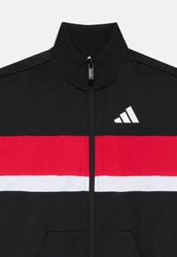 Svart zip-up jacka med hög krage; har djärva röda och vita horisontella ränder, texturerad ribbad krage och en vit logotyp.