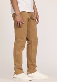 Chino en coton beige avec une coupe slim, dotés de coutures latérales et d'une texture lisse, associés à des baskets blanches et une chemise blanche décontractée.