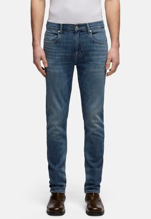 Uomo che indossa jeans blu slim fit e stivali di pelle marrone, in piedi con le mani rilassate lungo i fianchi davanti a uno sfondo neutro.