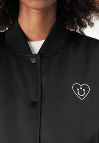 Zwarte bomberjack met ribbelkraag, voorzien van drukknoopsluiting en een witte hartlogo met de letters "HGOU" erin. Glad textiel.